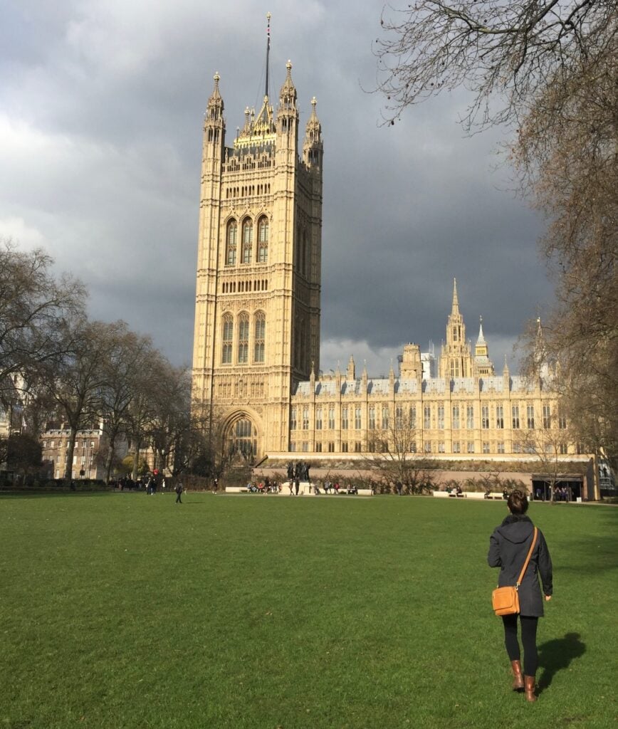 A person walks across a green lawn towards the towering Victoria Tower, part of the Palace of Westminster in London, with a dramatic sky overhead. The golden accents of the Gothic architecture contrast against the dark clouds.