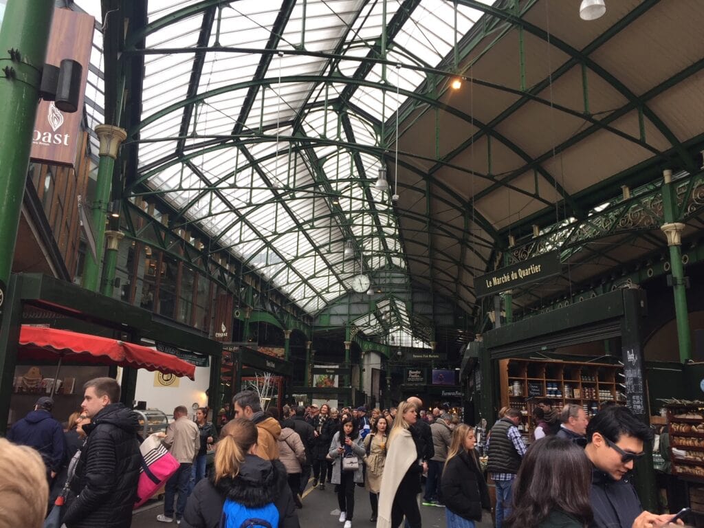 A crowded indoor market with high, green iron beams supporting a glass ceiling. Shoppers walk among stalls, including one with a "Le Marché du Quartier" sign, and others selling goods like bread. The setting suggests a lively atmosphere in an old market hall.