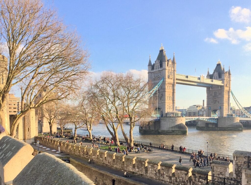 A scenic view of London's iconic Tower Bridge on a bright, sunny day. The bridge spans the River Thames, framed by leafless winter trees and a busy riverside with tourists walking along the path.