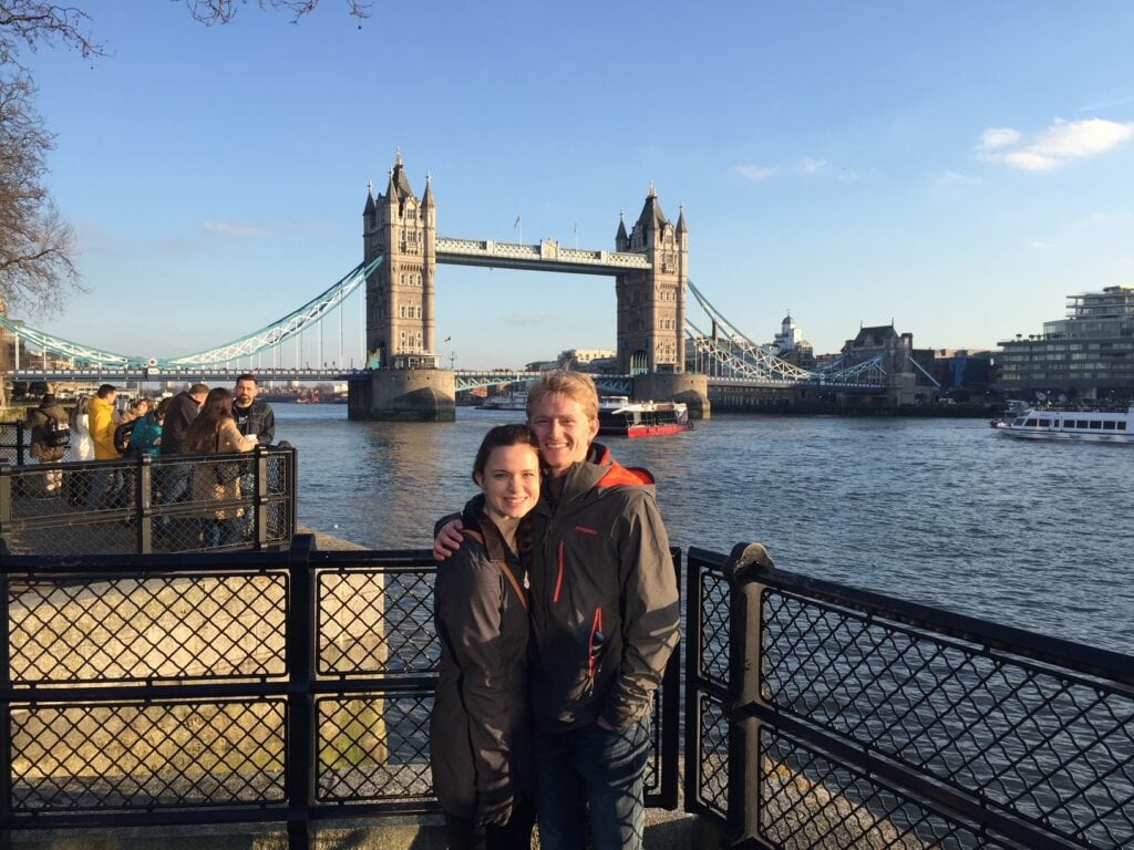 A smiling couple stands by the riverbank with the iconic Tower Bridge in the background on a sunny day. The man has his arm around the woman as they pose in front of the Thames River. Boats can be seen passing under the bridge.