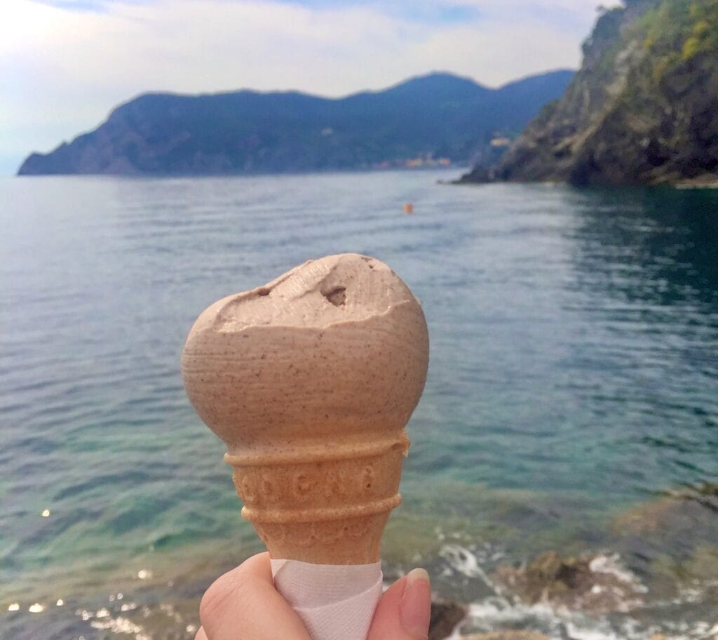 A close-up of a hand holding a scoop of cinammon gelato in a cone, with a backdrop of calm, clear water and distant mountains. The serene ocean and green hills create a peaceful, scenic view.