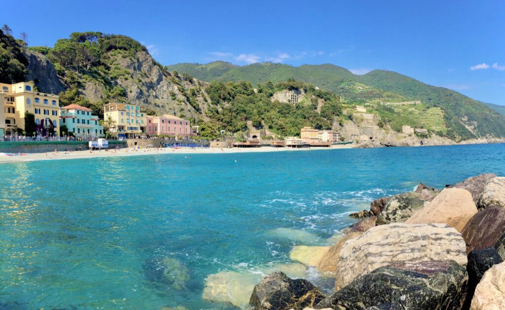 A scenic beach with turquoise waters gently lapping against large, rocky boulders in the foreground. Colorful buildings line the coast, with a backdrop of lush green hills. The bright blue sky and sun-dappled sea create a vibrant and serene atmosphere.