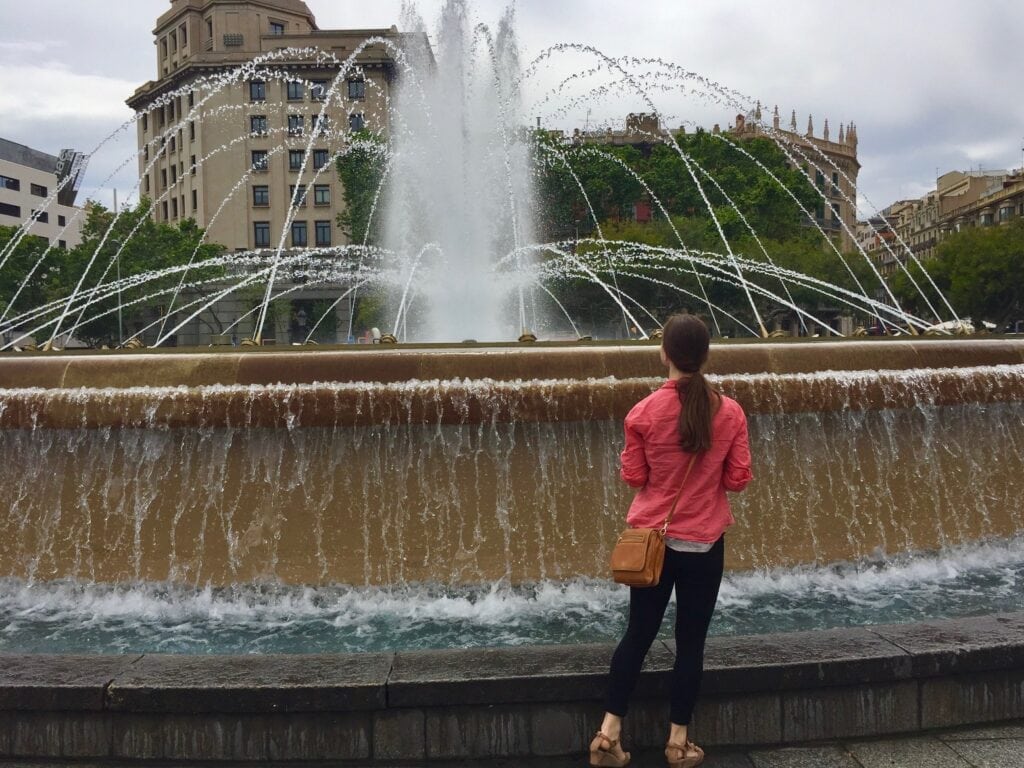 A woman, seen from behind, stands in front of a large fountain in a public square, with water shooting in arcs. She is wearing a pink jacket and black pants, and a tan purse hangs by her side. In the background, there are trees and classic European-style buildings.