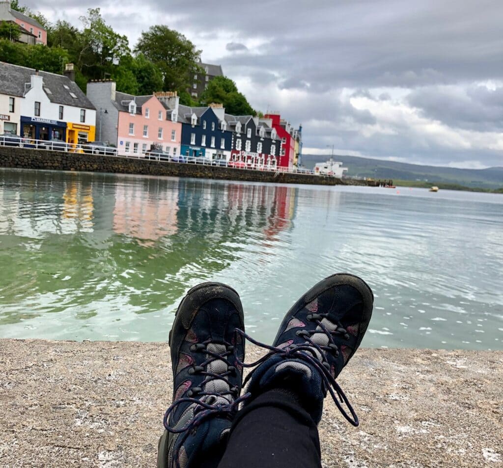 A pair of hiking boots are in the foreground, with colorful waterfront houses reflecting in the calm water. The scene appears tranquil, with overcast skies and picturesque houses painted in pastel colors along the harbor.