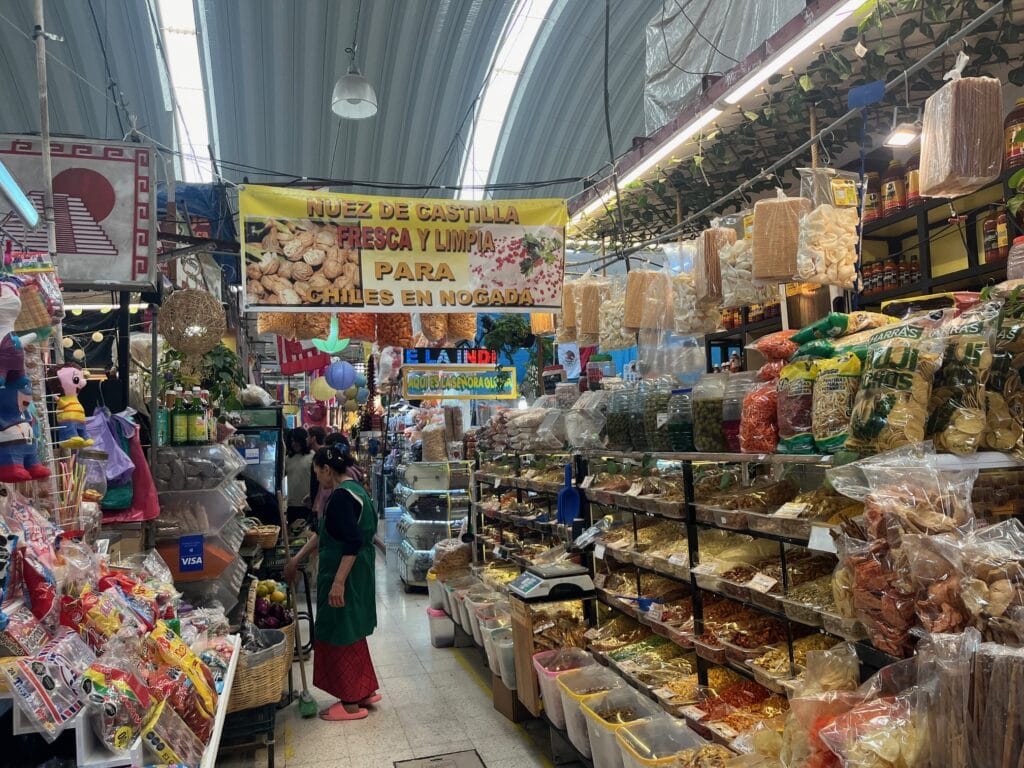 A bustling indoor market with shelves stacked high with various dried goods and snacks in plastic bags. A vendor dressed in a green apron tends to her stall, while a banner overhead advertises "Nuez de Castilla" for chiles en nogada. The scene is colorful and vibrant with a variety of packaged foods displayed.