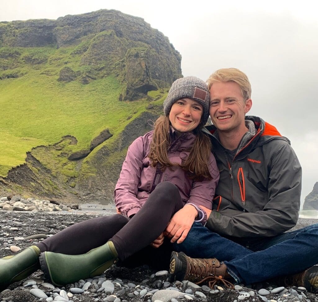 A couple sits on a black pebble beach, leaning against each other, smiling. Behind them, green cliffs rise into the misty sky, adding a dramatic backdrop to this cozy outdoor moment. Both are dressed warmly in jackets and boots.