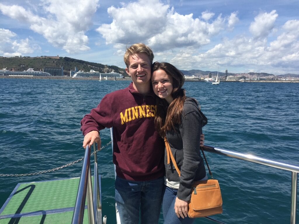 A young couple is smiling and posing on a boat with a scenic ocean backdrop. The man is wearing a "Minnesota" sweatshirt, while the woman leans in close, holding a small tan purse. Sailboats are visible on the water, and hills with buildings are in the background under a partly cloudy sky.