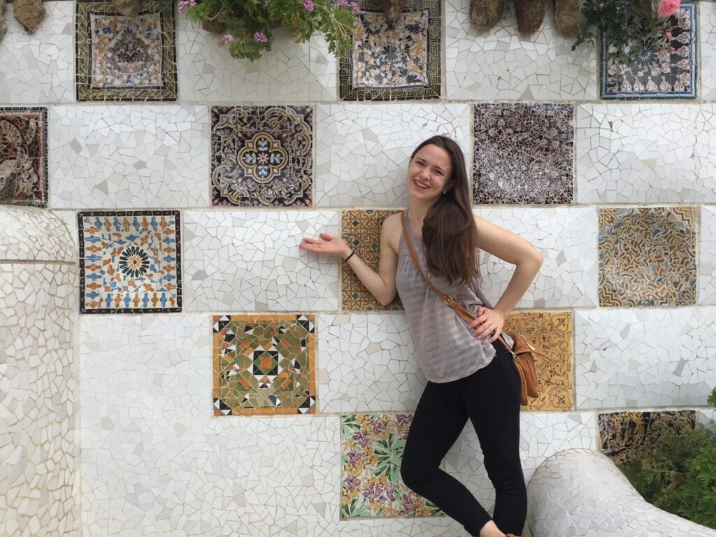 A woman smiles while standing in front of a colorful tiled wall with intricate mosaic designs. She is gesturing towards the wall with one hand and holding a tan purse, wearing a casual outfit of a sleeveless top and black pants.