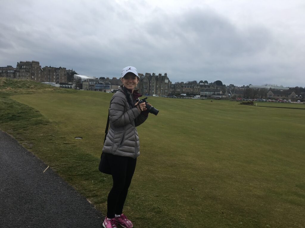 A woman stands on Old Course in St. Andrews Scotland, smiling at the camera while holding a DSLR camera. She is wearing a gray jacket, black pants, pink sneakers, and a white baseball cap. The background shows historic stone buildings under a cloudy sky.