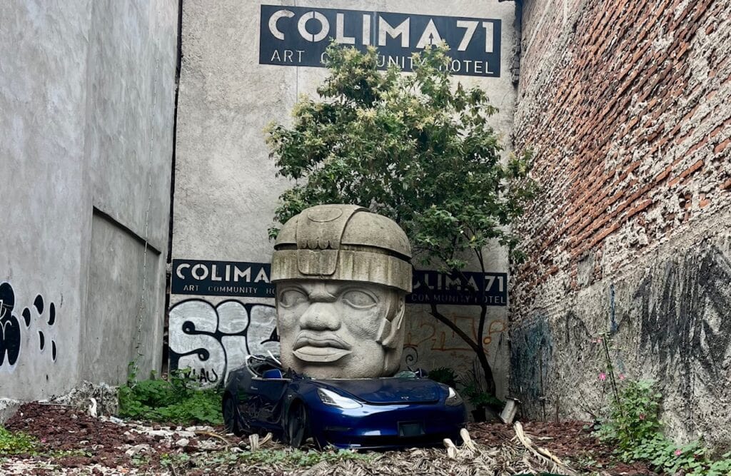 A large Olmec-style stone head sculpture sits on top of a blue car in a narrow alley, flanked by weathered walls with graffiti and signage for "Colima 71 Art Community Hotel." Overgrown plants surround the scene, giving it a surreal appearance.