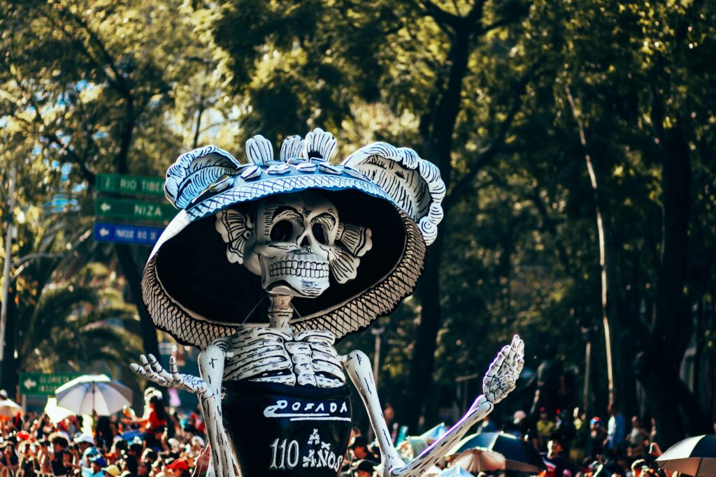 A towering skeletal figure of La Catrina, an iconic symbol of Día de los Muertos, dressed in a traditional hat with feathers, stands in front of a crowd under tree-lined streets. The costume bears the name "Posada" and the number "110 años," referencing the centennial celebration of the famous Mexican artist's creation.