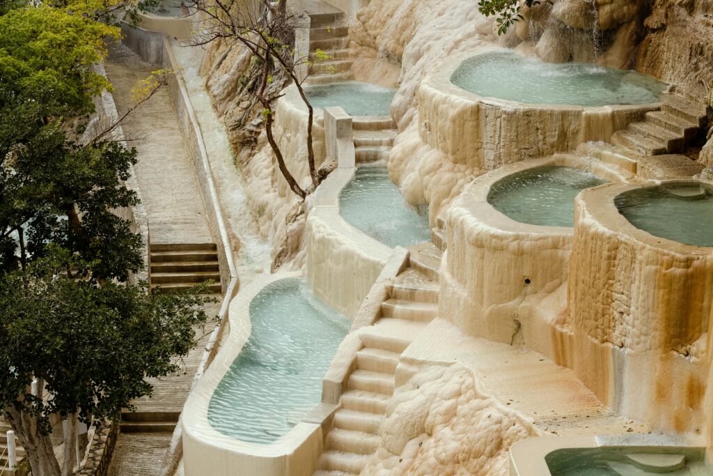  A series of natural hot spring pools cascade down a terraced, rocky hillside. Each pool is filled with turquoise water, separated by stone steps and ledges, with greenery and tree branches overhanging the tranquil setting.